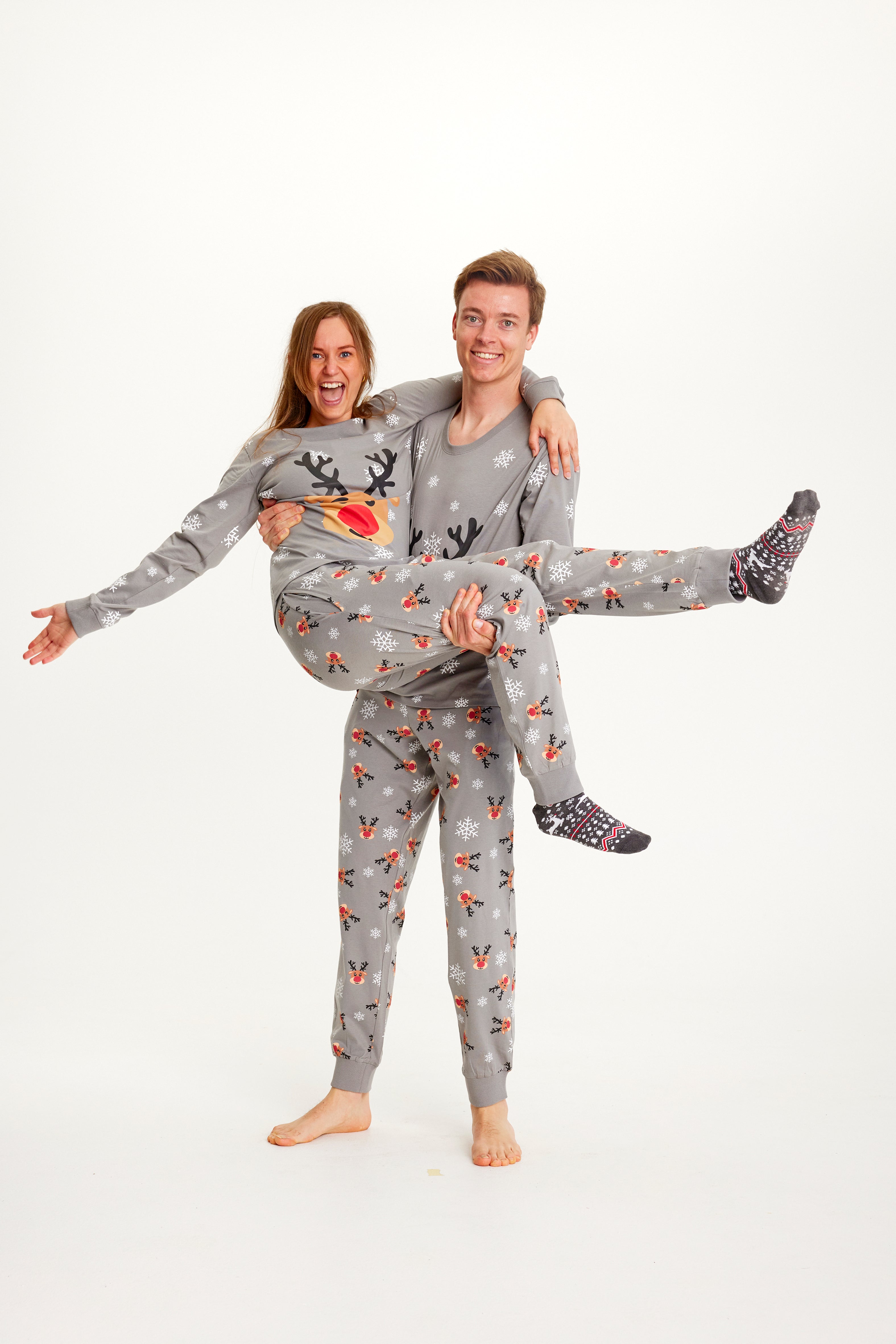 En mann som bærer en kvinne i grå julepyjamas med reinsdyr og snøfnugg.